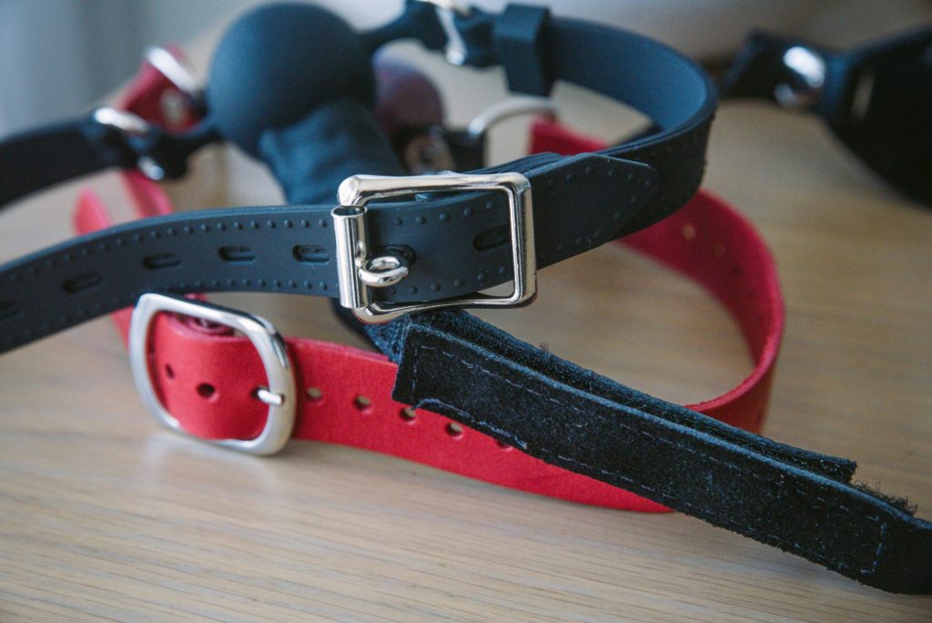 Two fastening straps from BDSM gags. One strap, a red one, has a traditional, normal fastening buckle. Another strap, a black one, has a locking buckle with no lock attached. 
