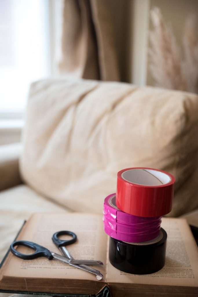 How to Use Bondage Tape. Image shows three rolls of bondage tape (in red, pink, and black) piled on top of a book next to a set of safety shears. 