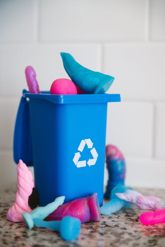 A blue recycling bin with the chasing-arrows, recycling arrow is overflowing with dildos. The dildos are overflowing the open top and are surrounding the base of the container. For how to recycle your sex toys article.