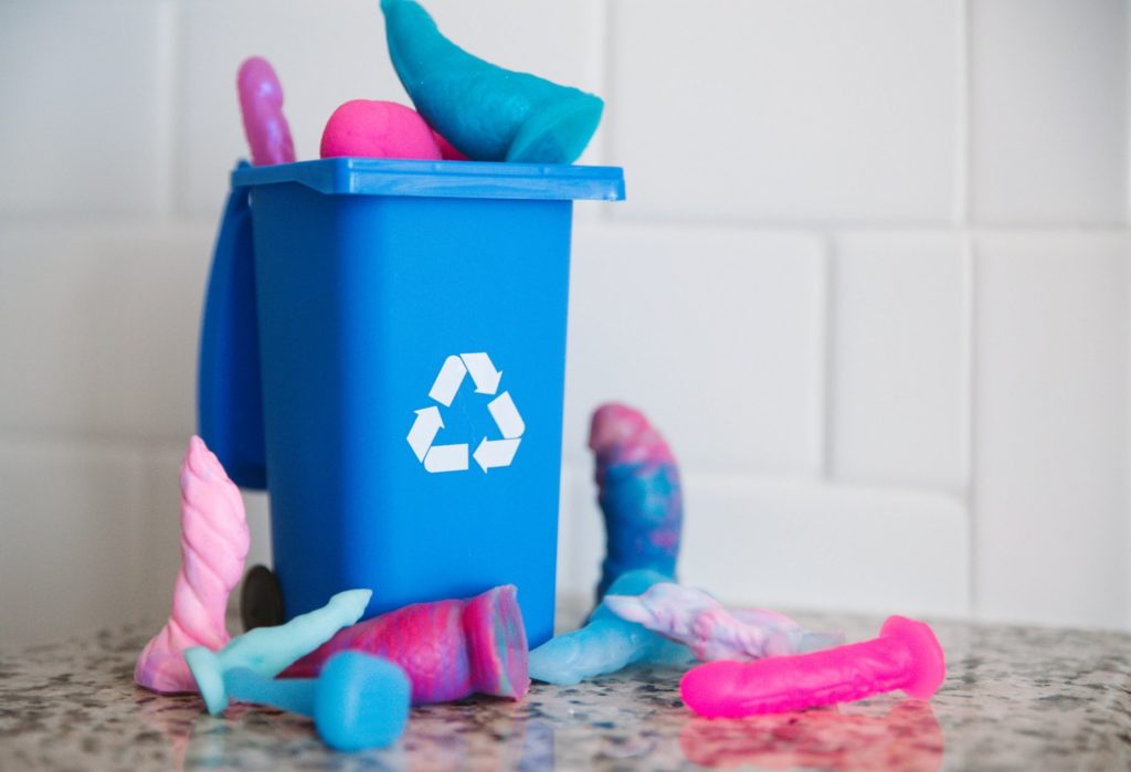 A blue recycling bin with the chasing-arrows, recycling arrow is overflowing with dildos. The dildos are overflowing the open top and are surrounding the base of the container. For how to recycle your sex toys article.