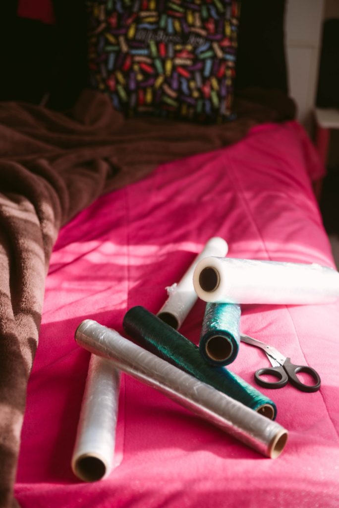 How to do Saran Wrap Mummification. Image shows rolls of colored saran wrap laying on the bed next to safety shears. Hard light makes it look like a calm, serene morning.