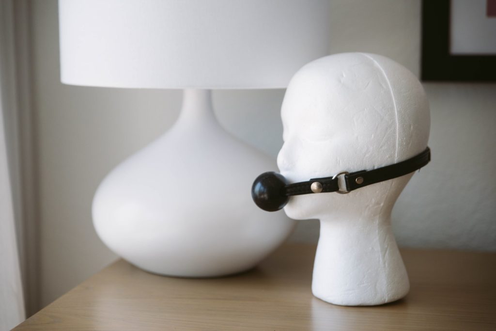 Styrofoam head on a table shows a mannequin wearing a BDSM ball gag. The bdsm gag is black with black leather straps, and the gag itself looks like a "ball" like the name implies.