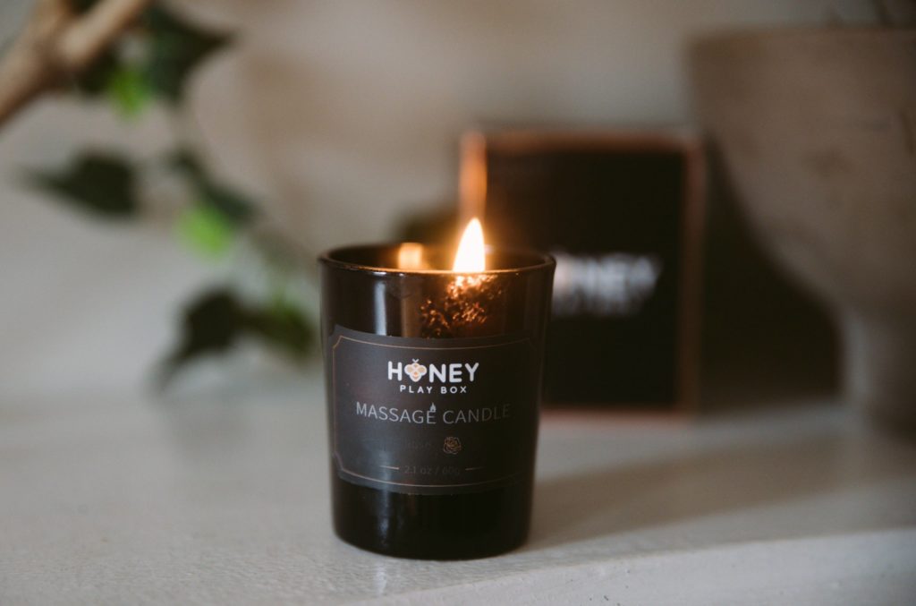 Honey Play Box Massage Candle in Rose. Image shows the candle burning in front of the massage candle's packaging. 
