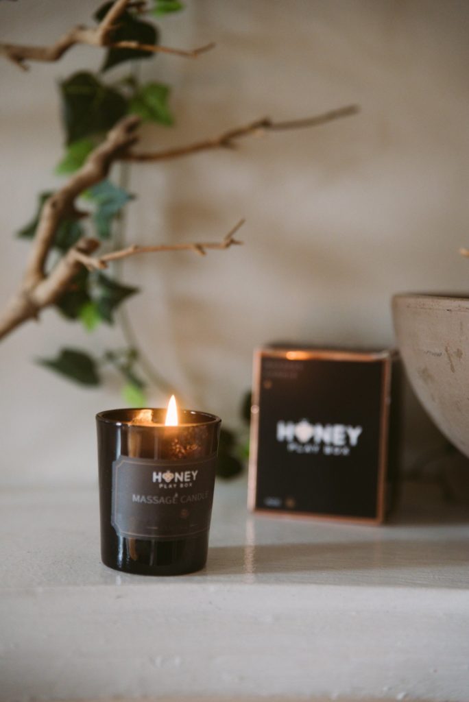Honey Play Box Massage Candle in Rose. Image shows the candle burning in front of the massage candle's packaging.