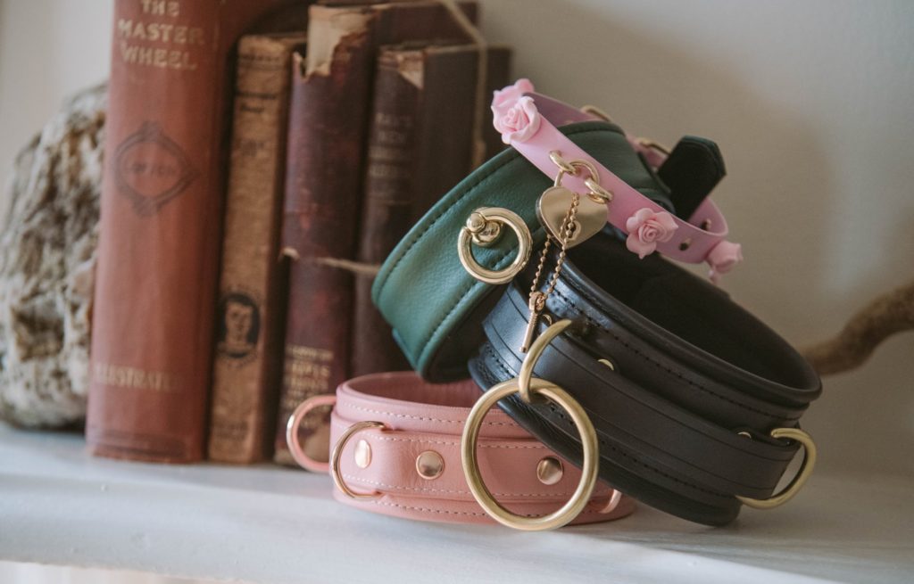 11 Tips for Choosing a BDSM Collar for Your Relationship. Image shows four separate bondage collars stacked on top of one another on top of a mantle next to some books.