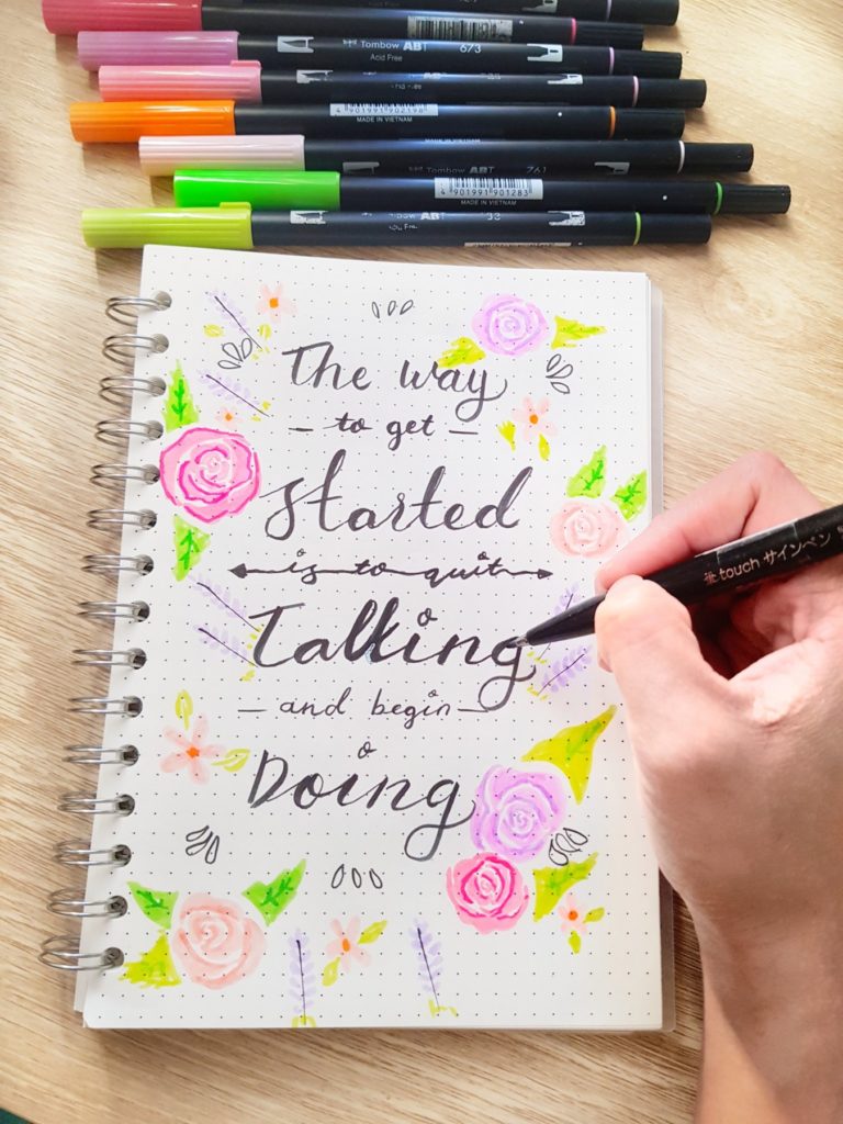 A notebook is shown with markers lined up on top of the notebook. The page says "The way to get started is to quit talking and begin doing" in pretty handwritten script. 