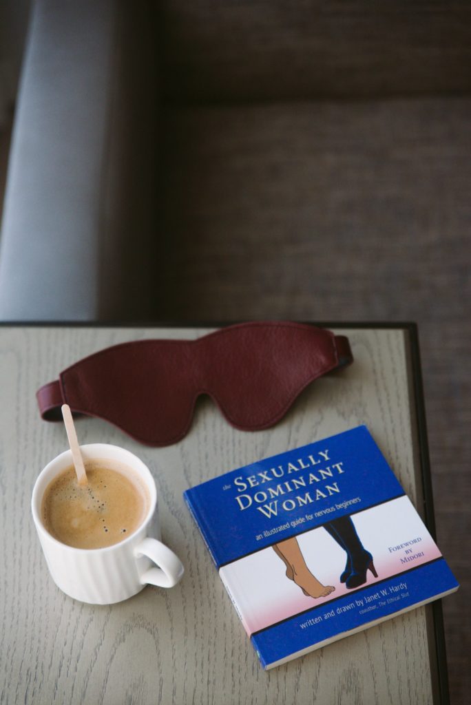 Sexually Dominant Woman Book Review. The book is sitting on a table next to a mug of coffee with milk and a wooden stir stick. A Liebe Seele dark red blindfold is shown in the background.