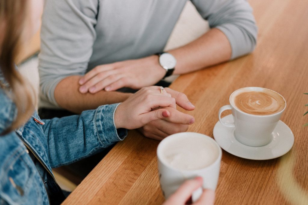5 Steps for Revealing Your Kinks to Your Partner. Image shows a couple sitting at a light brown table, both with coffee in hand. Their free hands are grasping in the middle of the table in a supportive gesture. 