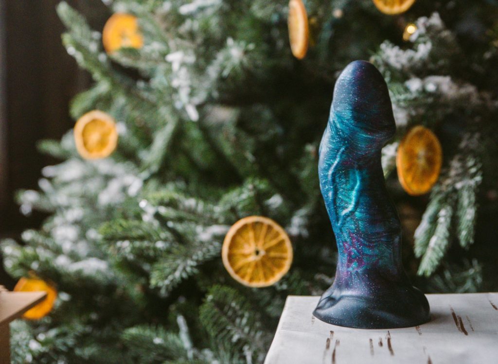 When You Shouldn't Buy Your Partner a Sex Toy image. Image shows a blue silicone dildo sitting out on top of a gift in wood-like wrapping paper. In the background, there's a Christmas tree with dried oranges being used as ornaments. 