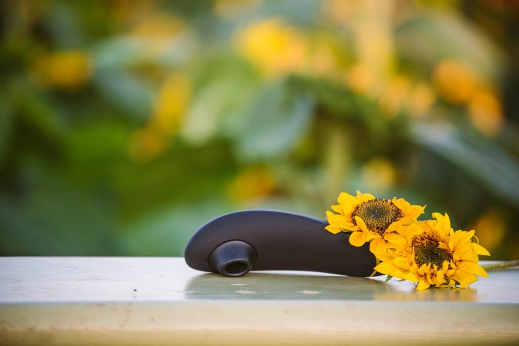 Womanizer Premium sitting out on a flat wooden surface outside. Along the handle, pretty sunflowers have been cut and placed. The image shows the blurry background of a field of sunflowers behind the wooden table the Womanizer Premium is on.