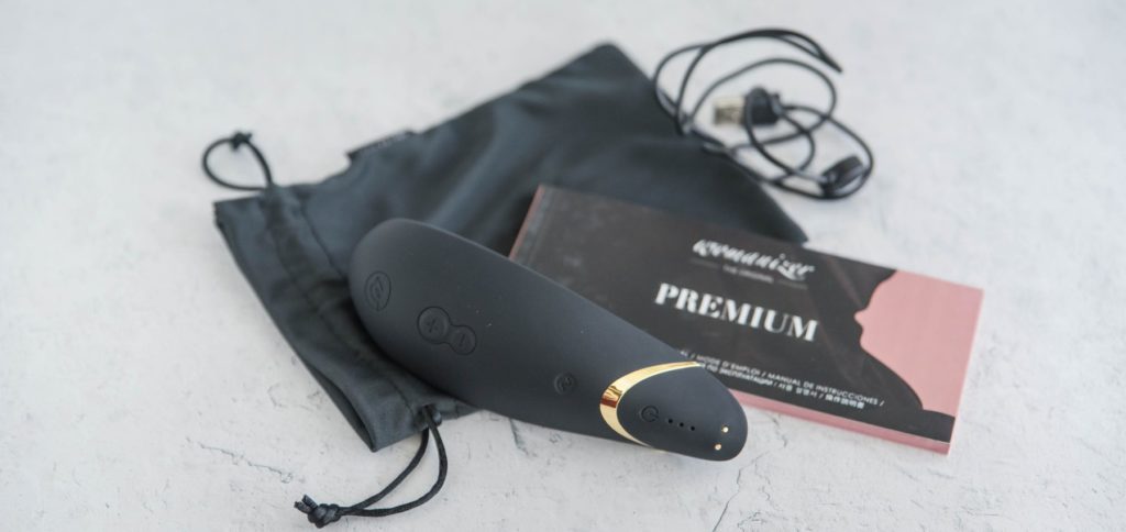 The Womanizer Premium laying out on a surface with everything that comes with the air suction vibrator. It has the rechargeable cord, the instruction booklet, and the drawstring storage bag.