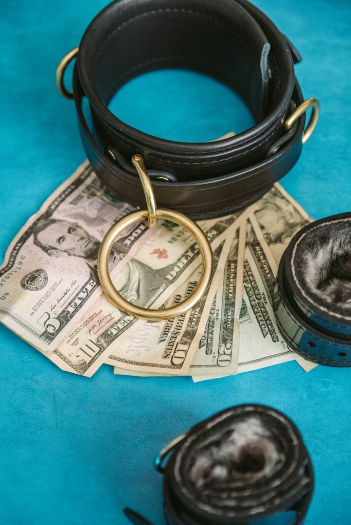 A BDSM collar and two cuffs sit out on a bright blue background. Underneath the collar, multiple denominations of US dollars are splayed out. Image for How to Explore a Money Kink as a couple.