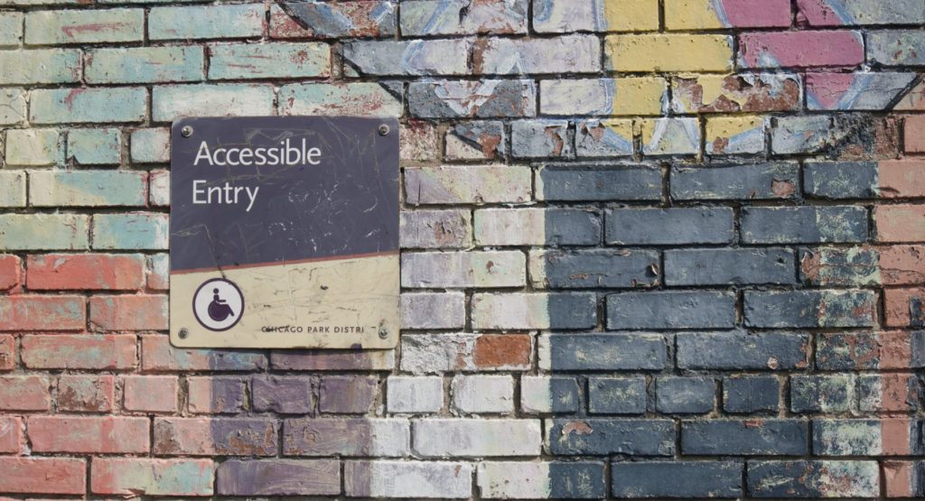 Decorative header image for accessible BDSM article shows a brick wall that looks like it may have been painted with graffiti. There is a close-up on an "Accessible Entry" sign for the Chicago Park District. 