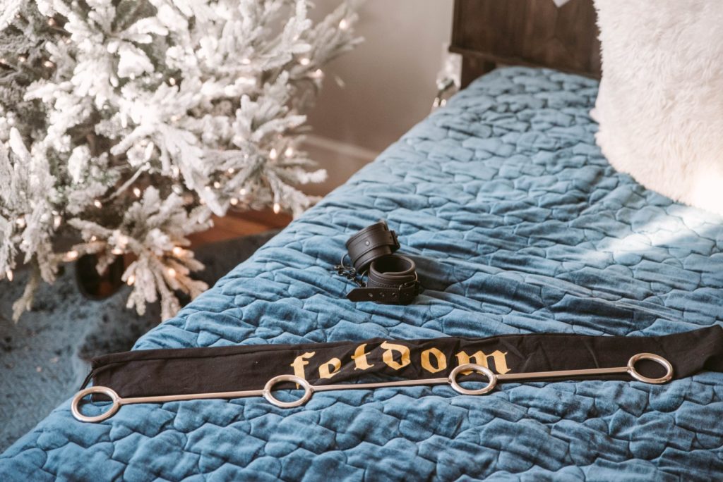 Fetdom Spreader Bar review. The Spreader Bar is sitting on a soft-looking bedspread on a bed. The spreader bar is sitting on top of its included, drawstring storage bag - and two of the four cuffs are sitting next to the spreader bar as well. In the background, a white artificial Christmas tree is shown with lights twinkling.