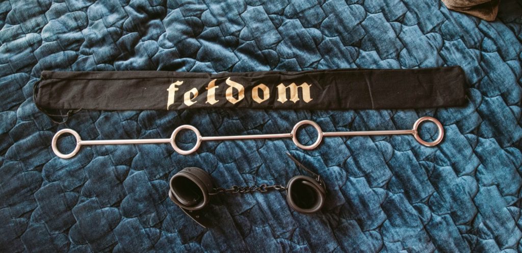 Fetdom Spreader Bar review. Spreader bar sits out on a blue, quilted bed spread. Above it is the long, black drawstring storage bag for storing the spreader bar. Two of the four cuffs are also shown next to the spreader bar.