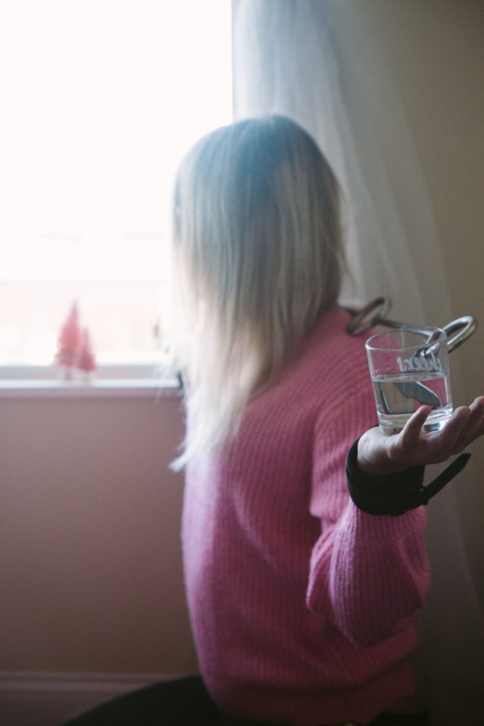 A person bound in the Fetdom Spreader bar in front of a window. They have long hair and are wearing a pink sweater. Their wrists are being held near their shoulders by the cuffs encircling their wrists that are attached to the spreader bar. In the center of each palm, there is a see-through glass filled with water. The glass is clearly slanted, and the water looks like it might tip soon.