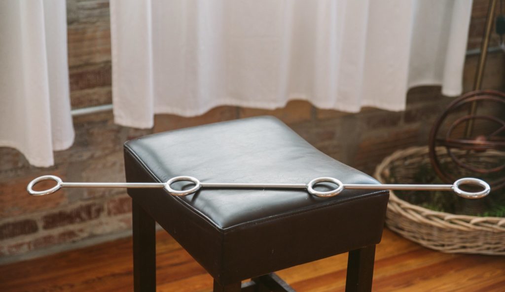 Fetdom Spreader Bar sitting on a dining stool. The ends of the spreader bar hang off the sides of the stool, but since the spreader bar is stainless steel, it doesn't bend or drop without the support of the stool.