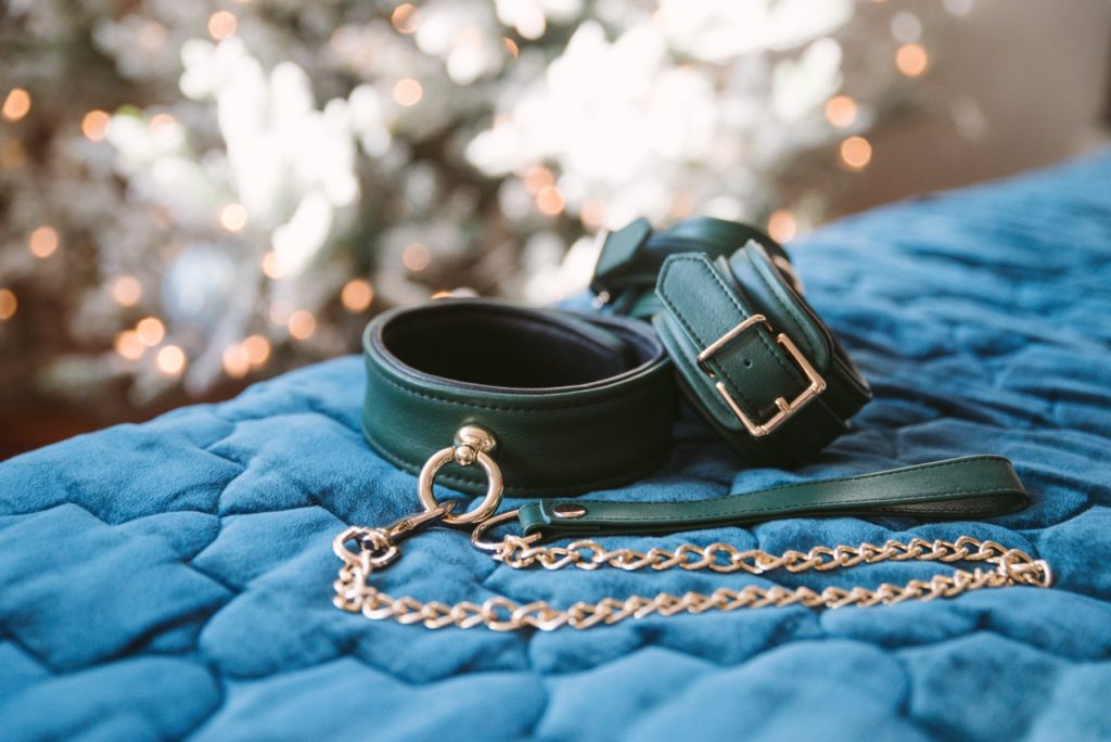 A set of green collar and cuffs with gold hardware from Liebe Seele sitting out on blue, quilted blanket. In the background, a white Christmas tree with lights can be seen. The image has an adorable, ethereal quality to it.