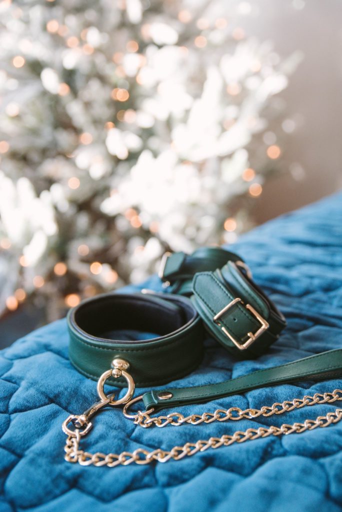 A set of green collar and cuffs with gold hardware from Liebe Seele sitting out on blue, quilted blanket. In the background, a white Christmas tree with lights can be seen. The image has an adorable, ethereal quality to it.
