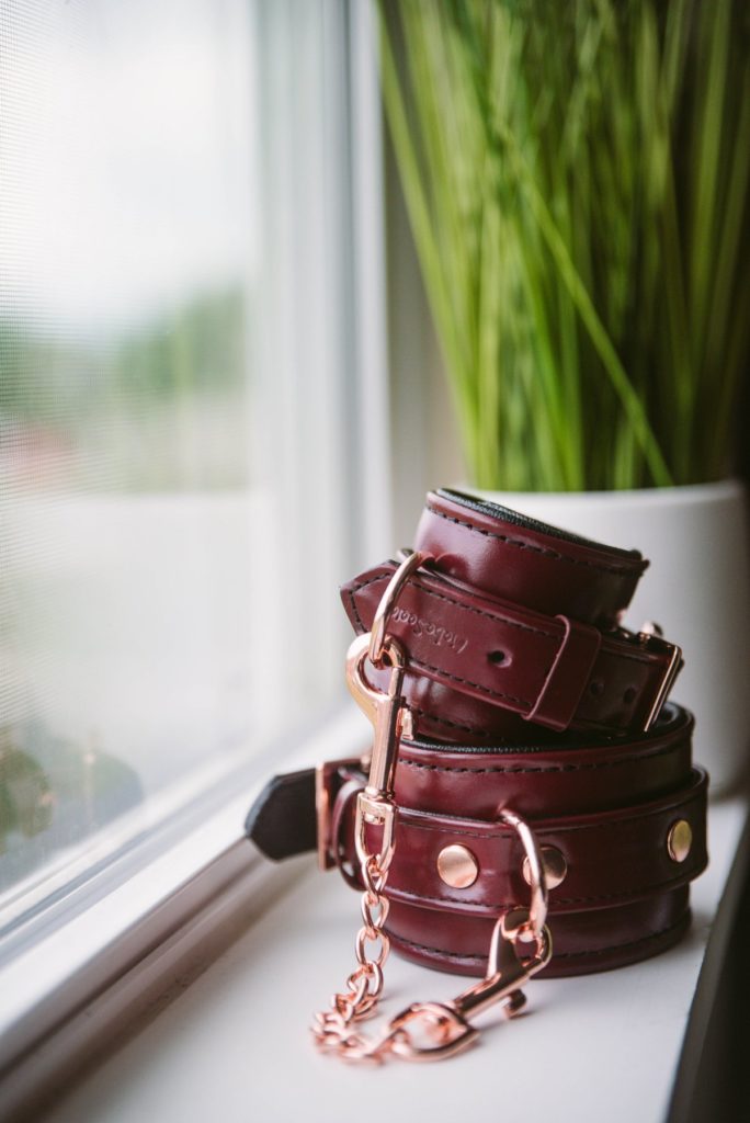 The Liebe Seele Wine Red Cuffs sit romantically on a windowsill with light, gentle light illuminating them. The rose gold and the deep wine red color really stand out. There's a pop of green color with a plant in the background.