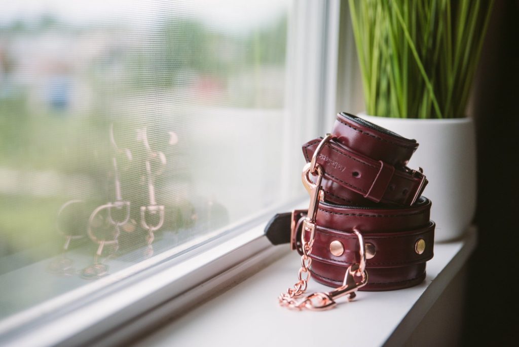 The Liebe Seele Wine Red Cuffs sit romantically on a windowsill with light, gentle light illuminating them. The rose gold and the deep wine red color really stand out. There's a pop of green color with a plant in the background.