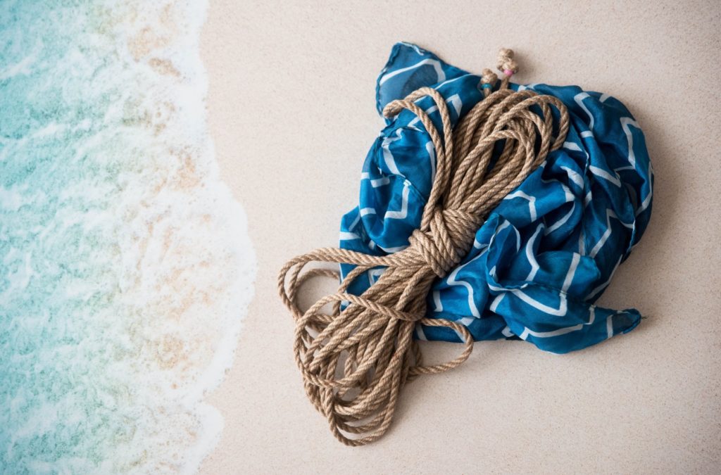 Image for my Water Bondage 101 article. A bundle of natural fiber rope is bundled up and sitting on top of a ocean blue scarf. The rope and scarf is sitting out on the sand with an ocean tide washing in.