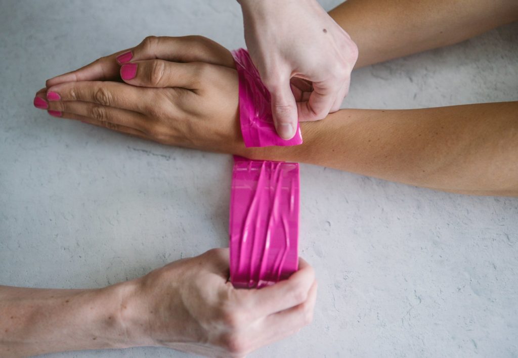 How to Use Bondage Tape image. A person's wrists are sitting out on a flat surface while another person's hands are wrapping bondage tape around the wrists. The pink bondage tape is pulled taut.