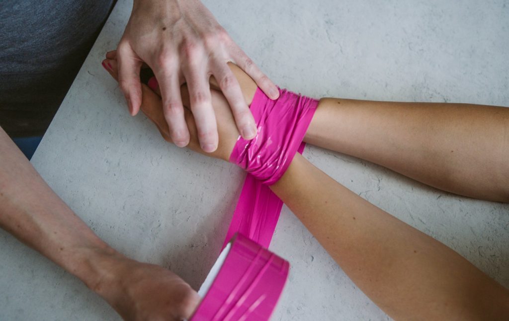 Two wrists shown side-by-side wrapped in pink bondage tape. A second set of hands is holding a roll of bondage tape and about to make another wrap around the wrists. Used for a how to use bondage tape article.