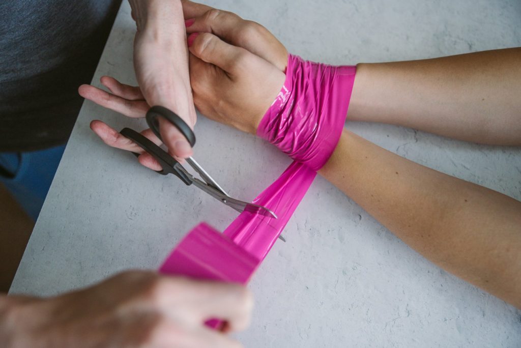 A person's two wrists are bound in bright pink bondage tape. Another person is cutting off a strip of the pink bondage tape with safety shears. Image for how to use bondage tape article.