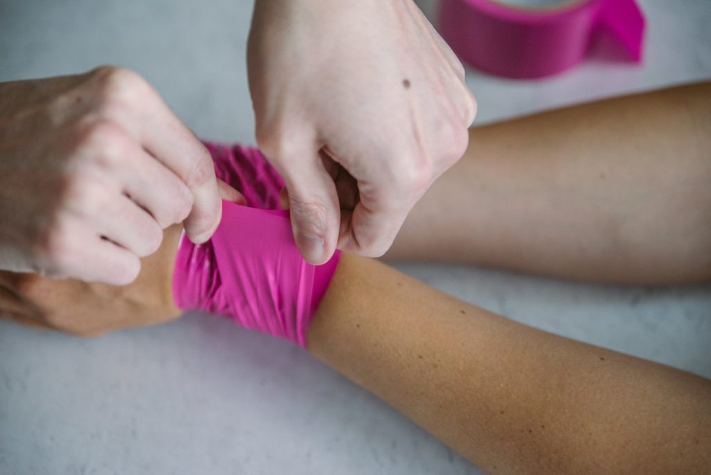 A person is holding the tail end of a piece of bondage tape. They are pressing it securely to a wrap of pink bondage tape around another person's wrists.