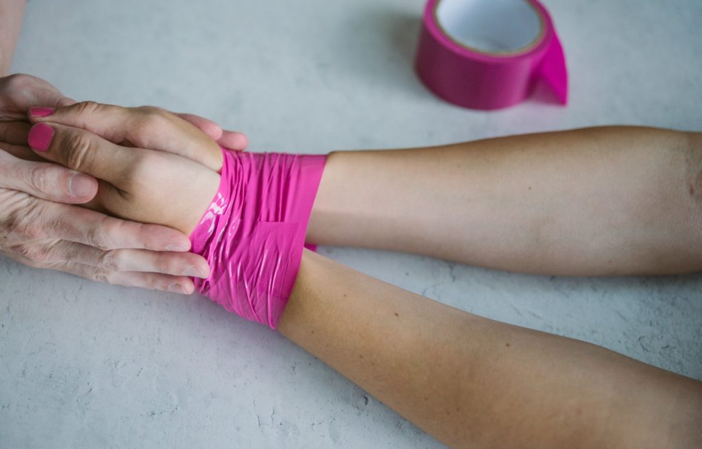 Two people hold hands in the middle of a white table. One person's wrists are bound in pink bondage tape.