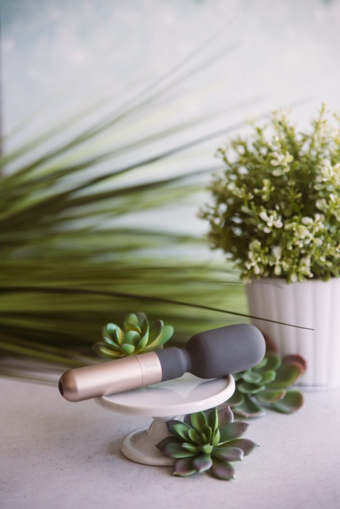 Love Not War Koi review: The sustainable wand massager sits on a white pedestal in front of a multitude of different types of greenery.