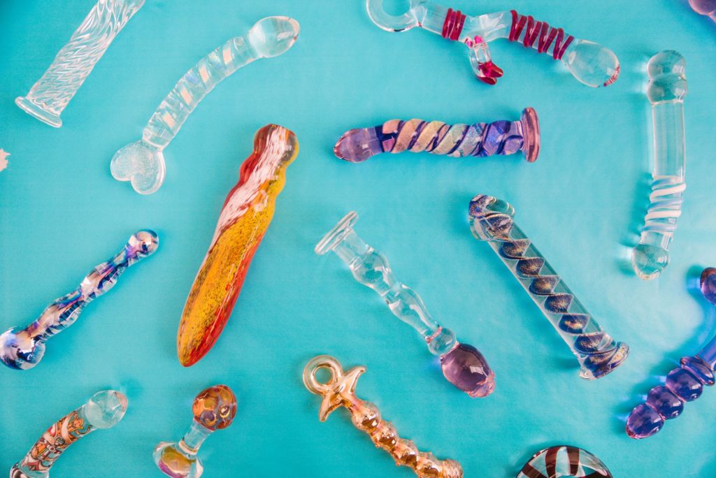 Why Use a Glass Dildo header image. A bunch of brightly colored, beautiful glass dildos are scattered out on top of a sky blue background. The image feels light and airy.