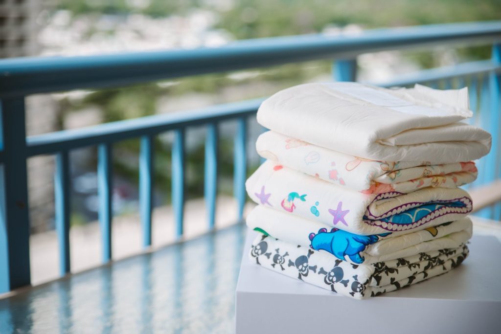 Pile of adult diapers sitting outside on a table on a bright and sunny day. The background shows that this table is sitting on top of a balcony. It all looks very colorful and approachable. For my Diaper Wearing Fetish 101 post.