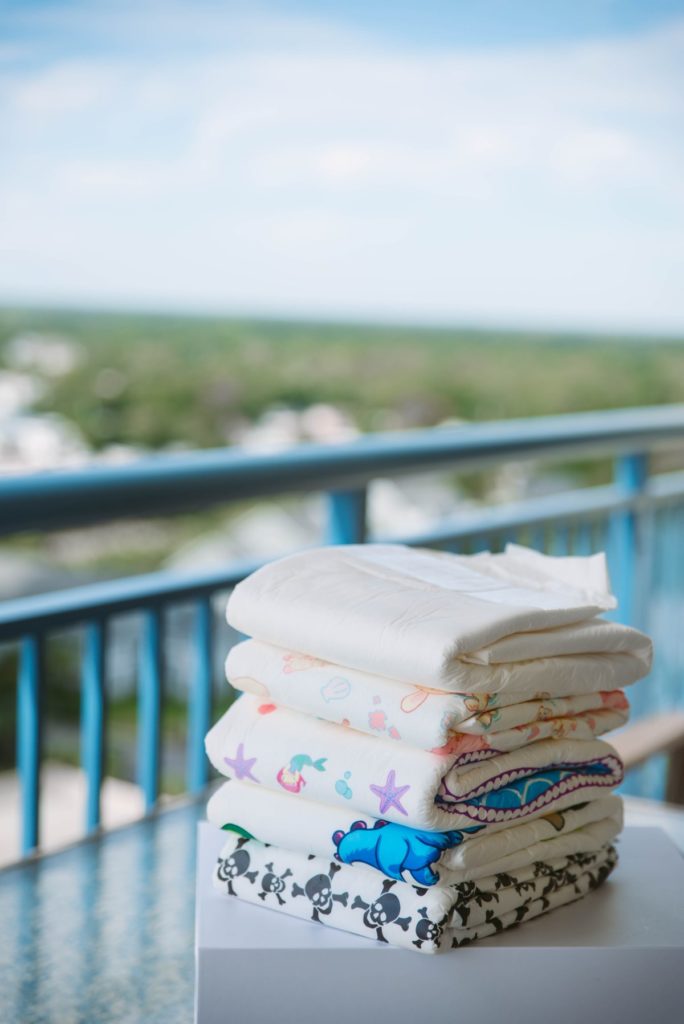 Pile of adult diapers sitting outside on a table on a bright and sunny day. The background shows that this table is sitting on top of a balcony. It all looks very colorful and approachable. For my Diaper Wearing Fetish 101 post.