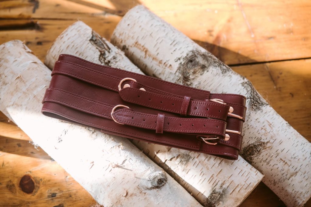 The Liebe Seele Wine Red Waist Cuff lays out flat on top of a few light wooden logs. The bright mid-day sun streams in around the Waist Cuff.