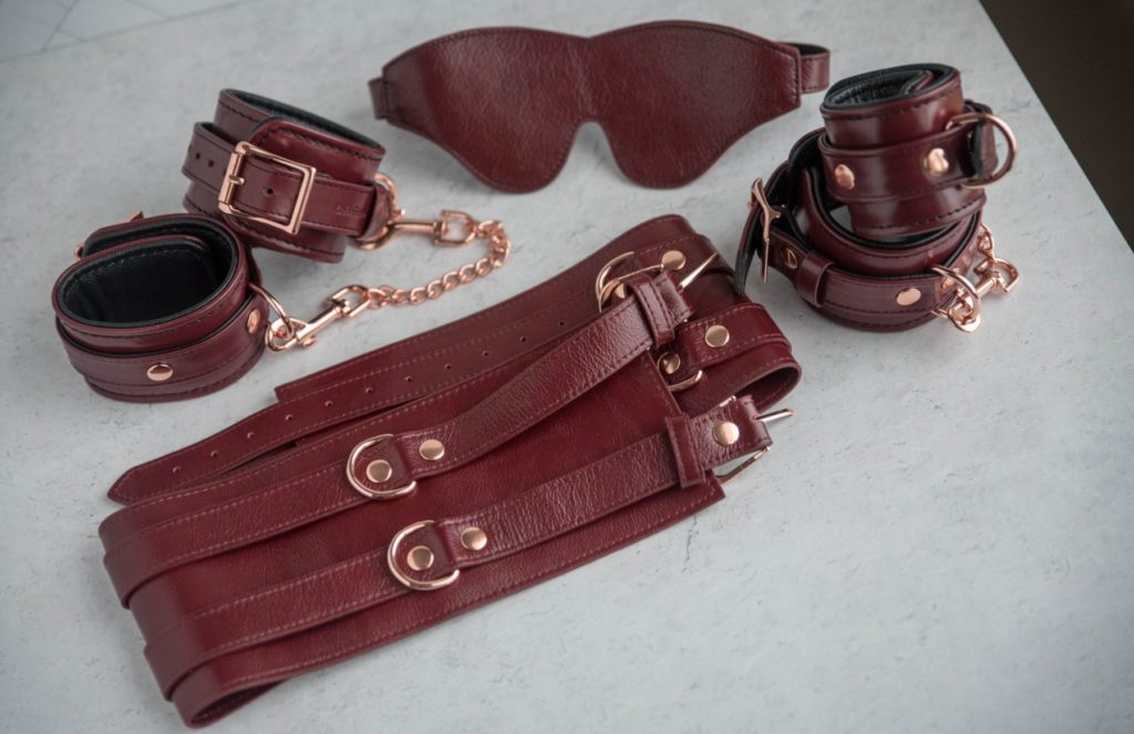 Liebe Seele Wine Red Waist Cuff Review: The Wine Red set sitting out on a surface. The blindfold, wrist cuffs, ankle cuffs, and waist cuff all match in style, hardware, and leather.