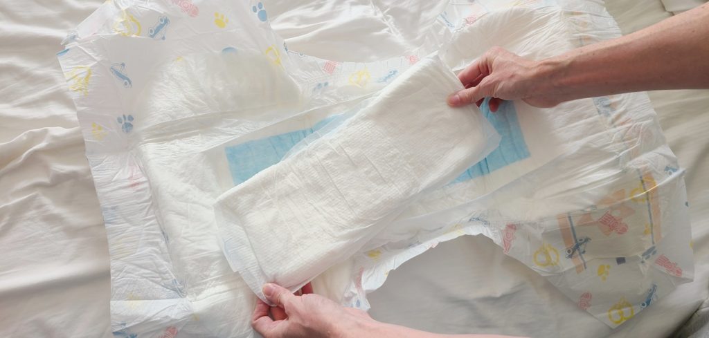 An adult diaper is opened and laid flat on a bed. Two hands are placing a stuffer/booster into the diaper to expand its capacity. The stuffer is very large compared to the diaper itself, and it covers up the entire crotch area of the diaper. 