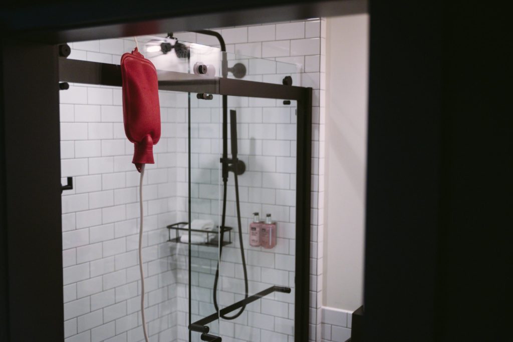 A enema bag hangs on top of a glass shower door. The entire bathroom is very white and tiled, and the red bag of the enema kit stands out. The white enema cord drapes out from the bottom of the bag. For my enema kink article. 