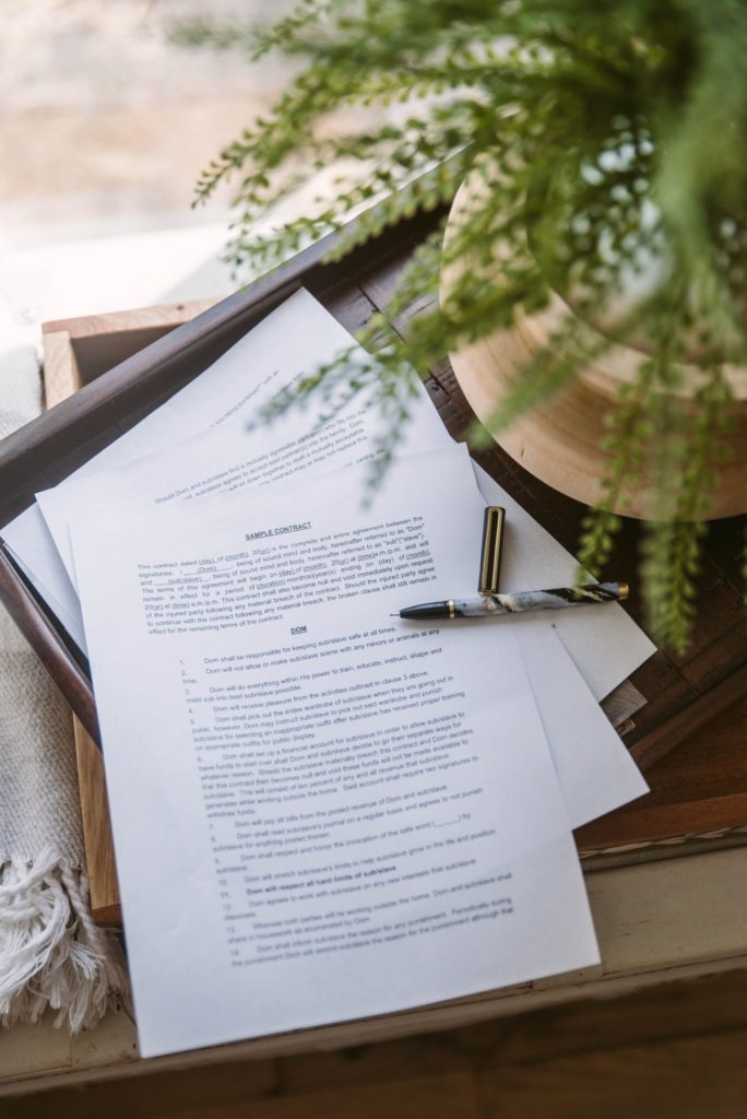 A BDSM contract shown sitting on top of a brown table with a black and gold pen open and laying next to it. A green plant partially obscures the contract. The entire image feels artsy. Image for my Protecting Yourself in Case of a D/s Break-up post.