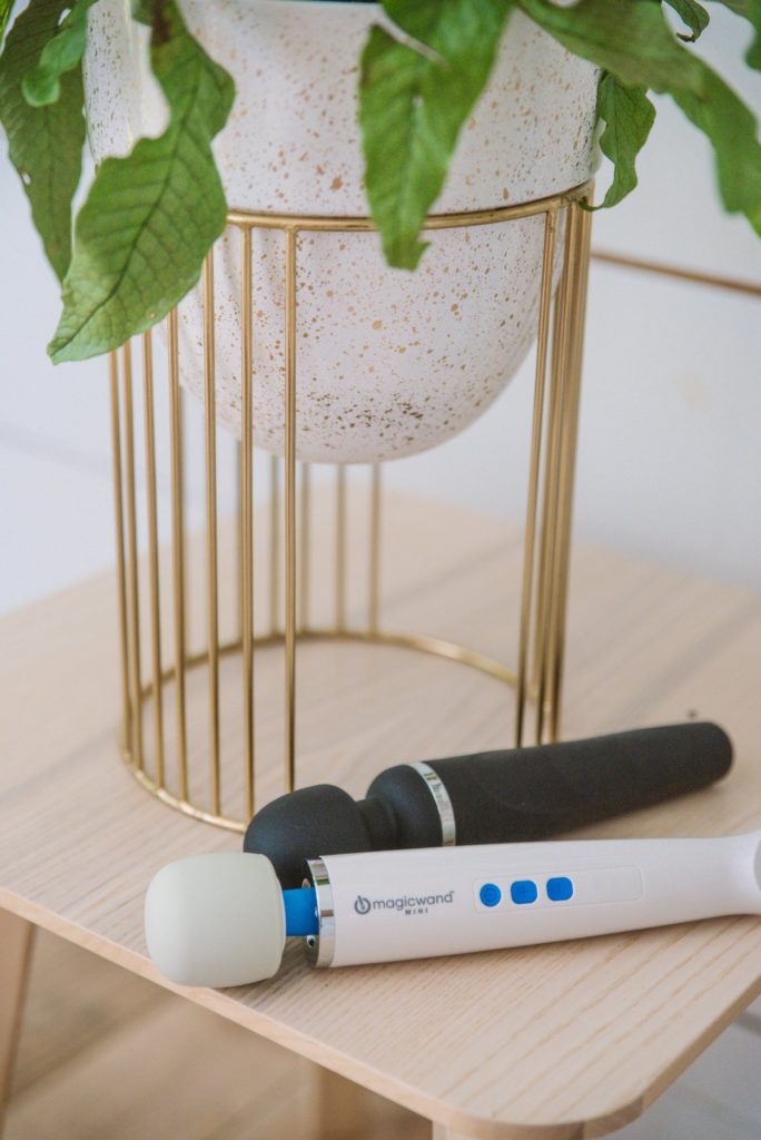 Magic Wand Mini vs Lovense Domi 2 Mini Wand Massager Comparison. Both of the wands are laying on a wooden bedside table next to the base of a plant in a geometric plant pot.