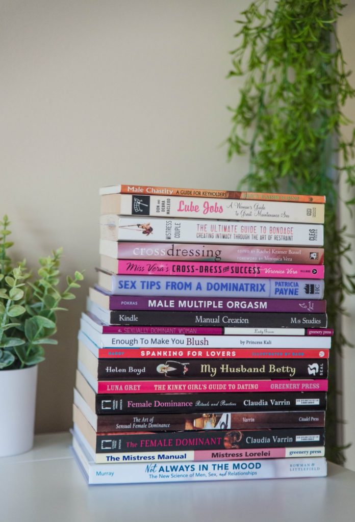 Best Domme Books to Introduce Femdom to your Wife image. A tall stack of femdom books are sitting on a white table in front of a bright green faux plant in the background.