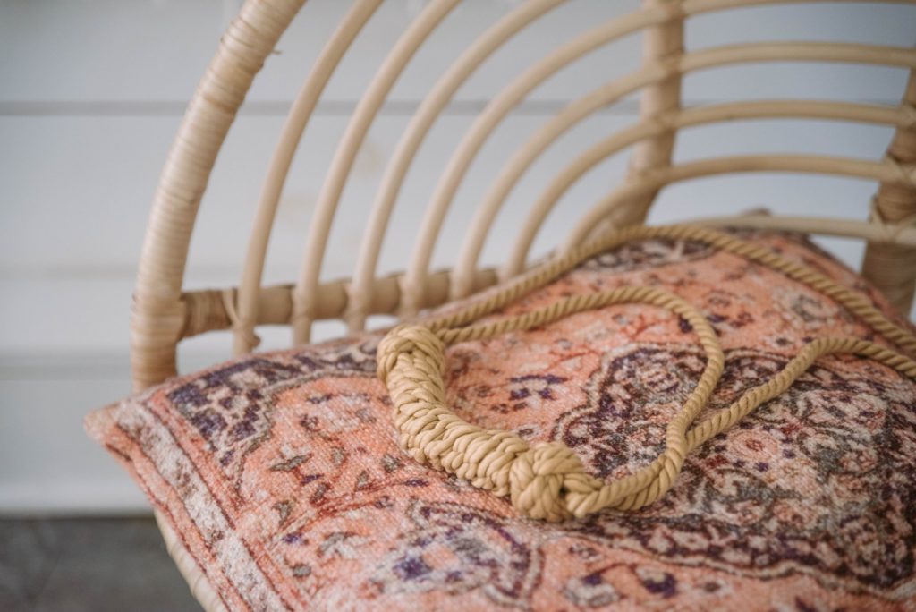 The Liebe Seele Bound You II shibari rope gag sits out on top of a wicker chair with a patterned cushion. The colors match almost perfectly, and it can be hard to make out the natural-colored rope of the gag from the wicker chair behind it. 
