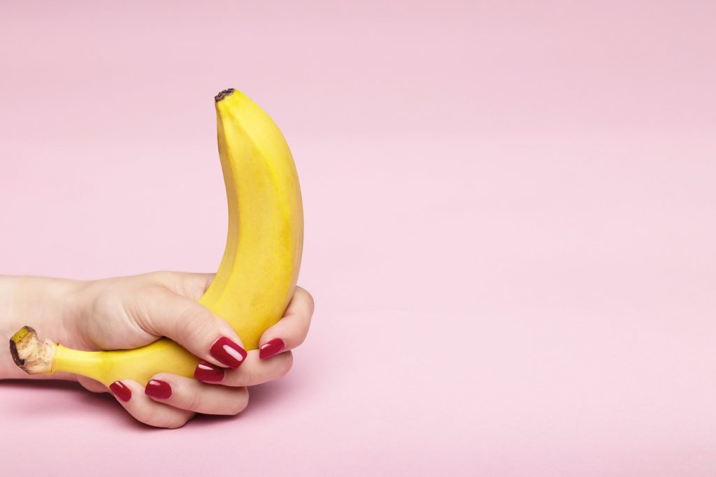 Display image for "how to give joi" article. A person with painted red fingernails grips a bright yellow banana in front of a pale pink background.