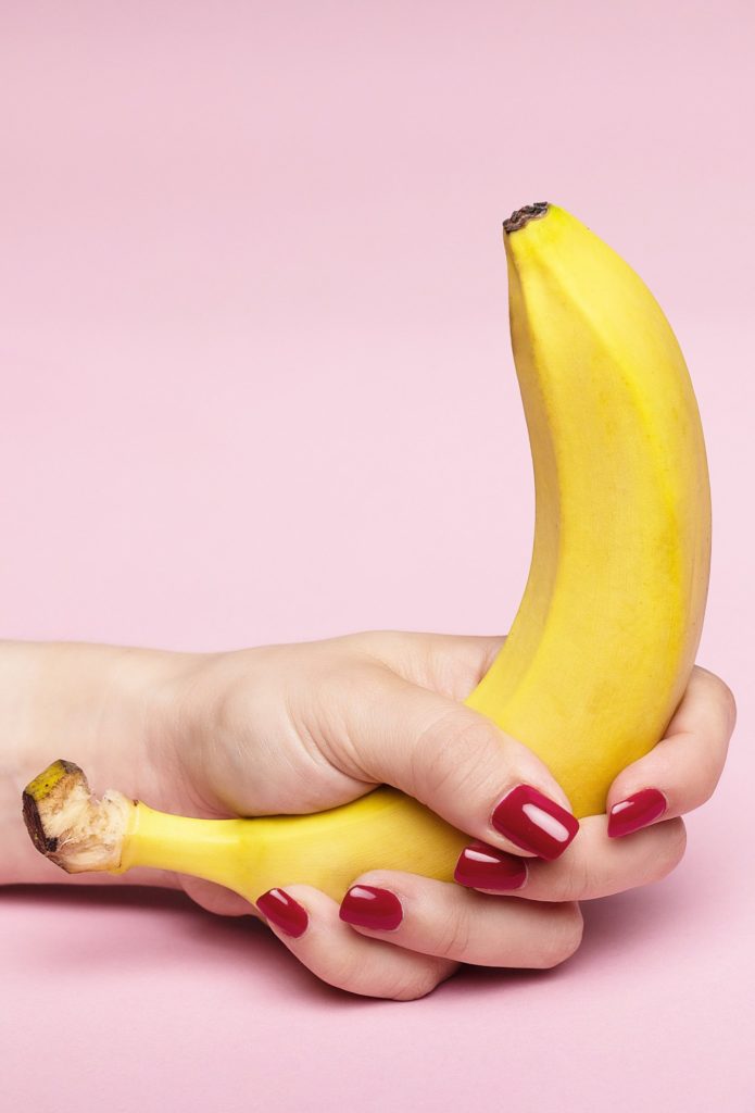 Display image for "how to give joi" article. A person with painted red fingernails grips a bright yellow banana in front of a pale pink background.