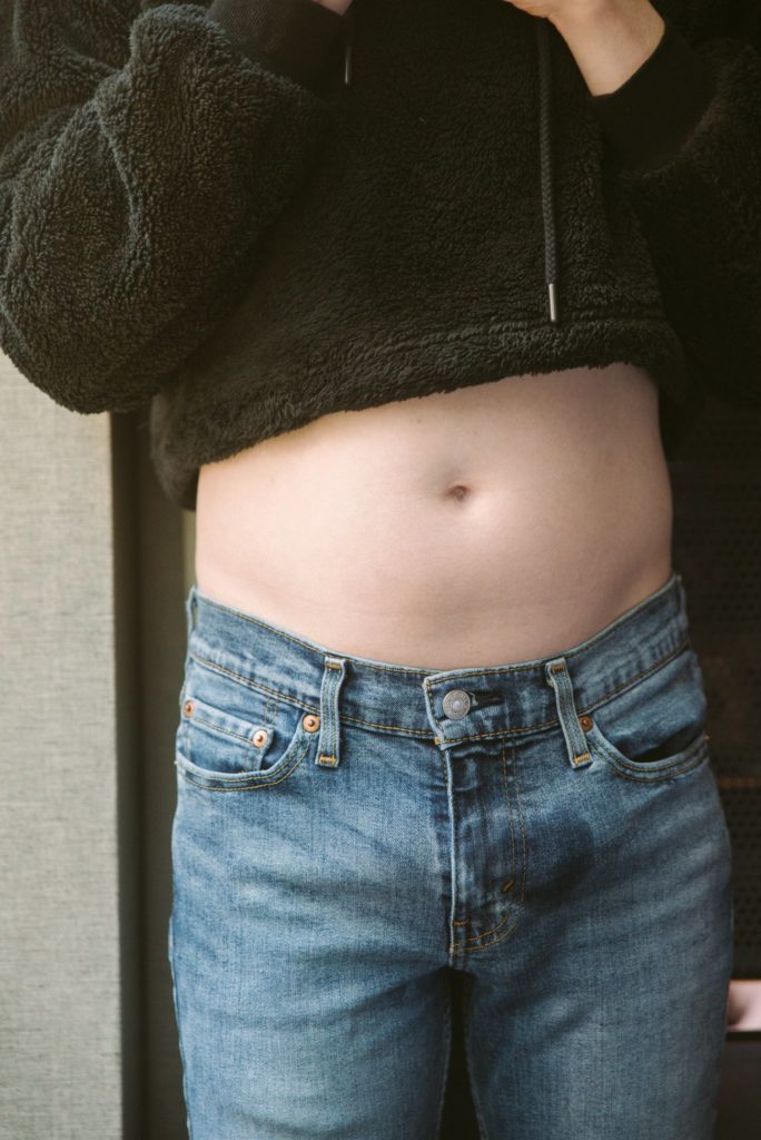 A person wearing snug denim jeans is standing, well-lit near a window. The soft skin of their abdomen is showing. There's a small, oval-like wet spot clearly visible on the front of the jeans.