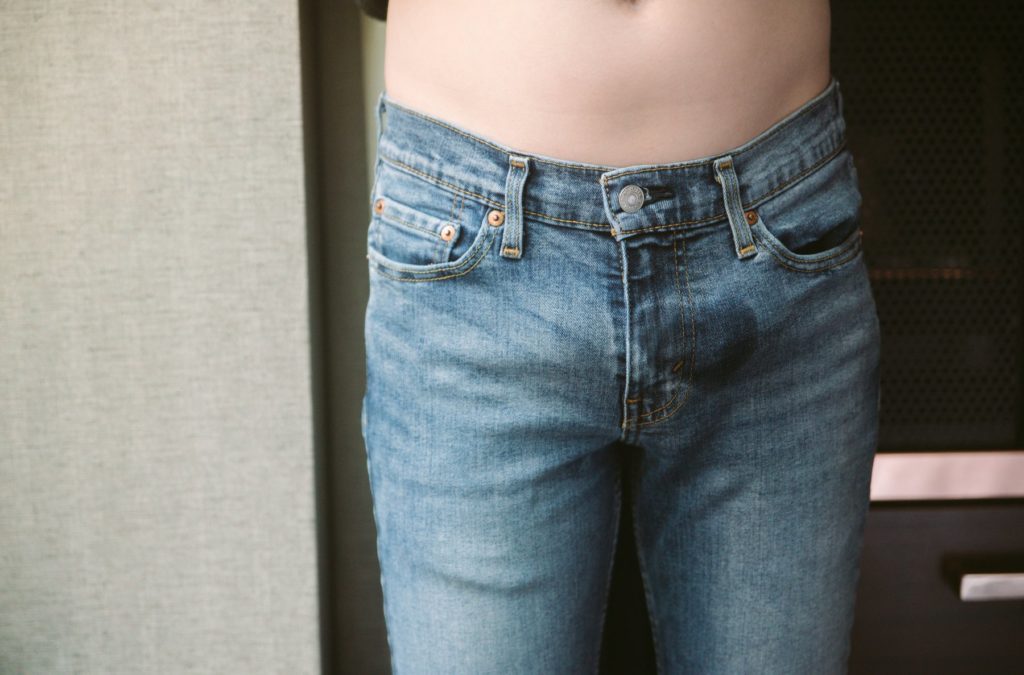 A person wearing snug denim jeans is standing, well-lit near a window. The soft skin of their abdomen is showing. There's a small, oval-like wet spot clearly visible on the front of the jeans.