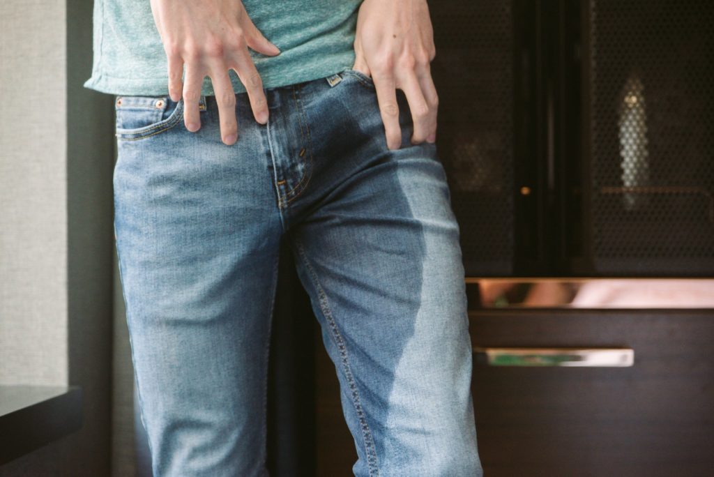 A person wearing snug denim jeans is standing, well-lit near a window. The soft skin of their abdomen is showing. The entire leg of one side of their jeans is dark blue and clearly wet. It looks like someone wet their pants.