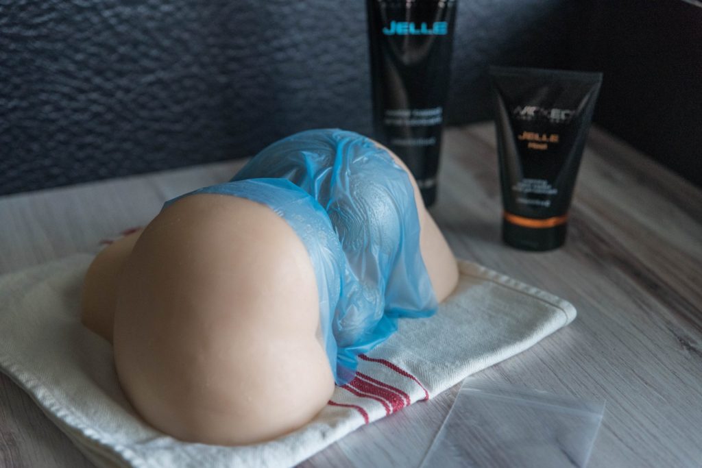 Image for How to Do Anal Rimming Safely. A miniature sex doll is laying on top of a wooden countertop. A dental dam is laying out over the anal area of the sex doll. Two bottles of Wicked Jelle anal lubricant are propped up in the background.
