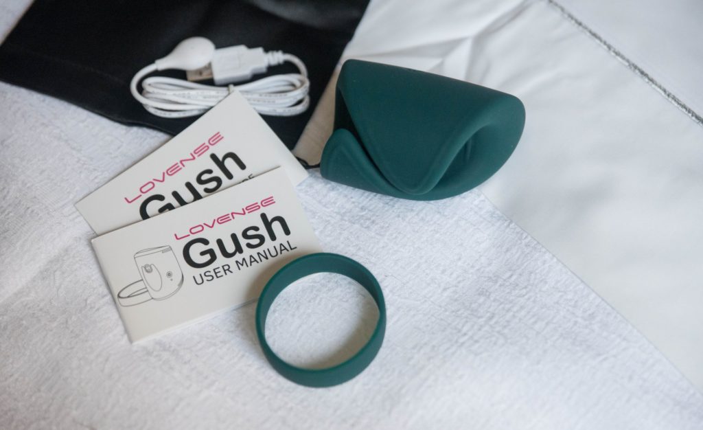 Everything that comes with the Lovense Gush laid out on the white bedding of a bed. There's the storage bag, the charging cable, instruction manuals, the Lovense Gush, and the optional bad that wraps around the Gush. For my Lovense Gush review.